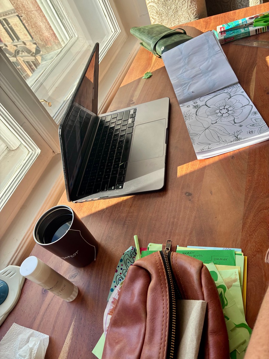 A laptop, sketchbook and art supplies on a sunny wooden desk by a window